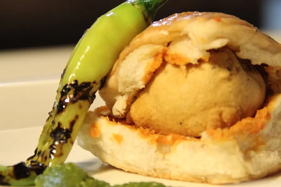 A vada pav in a soft bun with a fried green chilli beside it